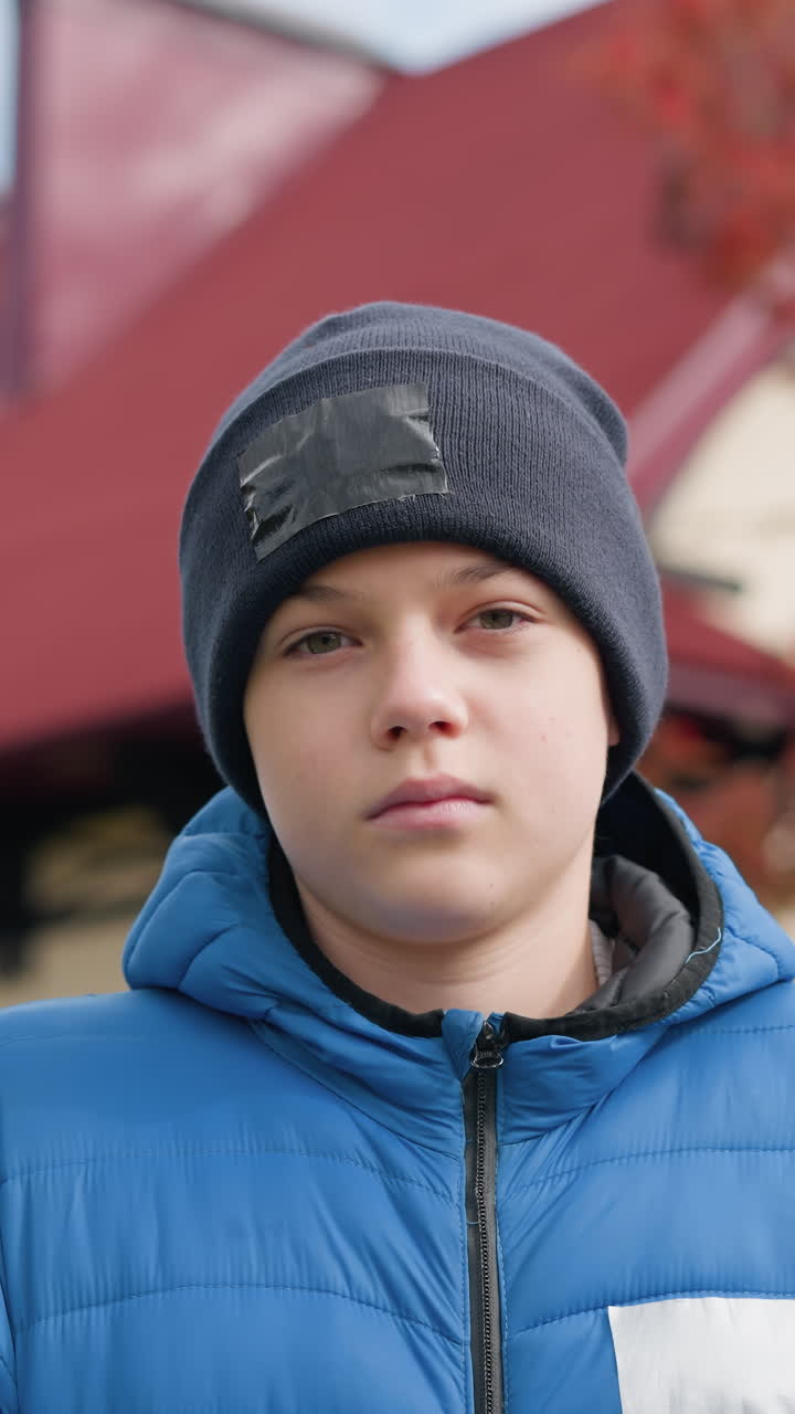 niño joven con gorra negra y chaqueta azul de pie al aire libre, mirando pensativo frente a hermosos árboles de otoño y casa con colorido follaje rojo en el fondo