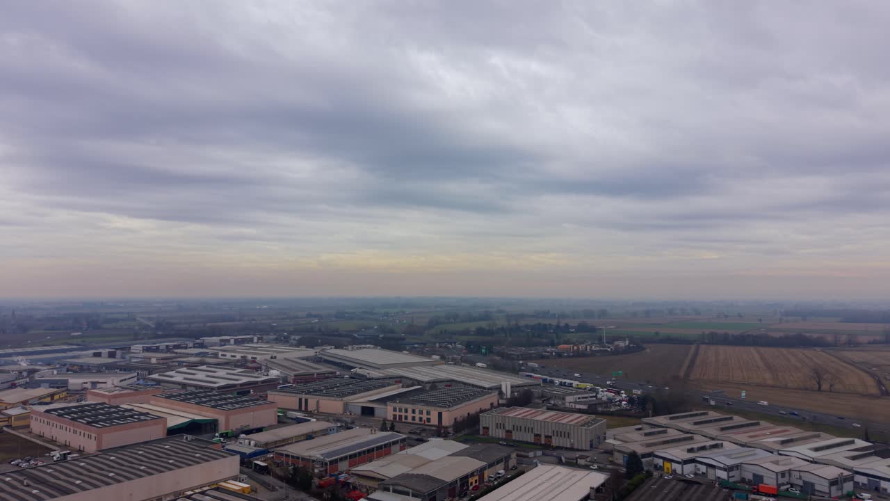 Filmato aereo panoramico che mostra un grande parco industriale con fabbriche e magazzini in una giornata cupa e nuvolosa. Un'autostrada trafficata costeggia la zona.