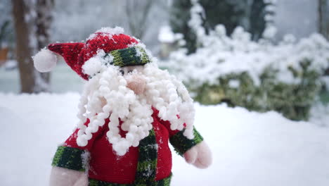 un juguete blando de papá noel está sentado en una mesa en un jardín mientras la nieve cae a su alrededor