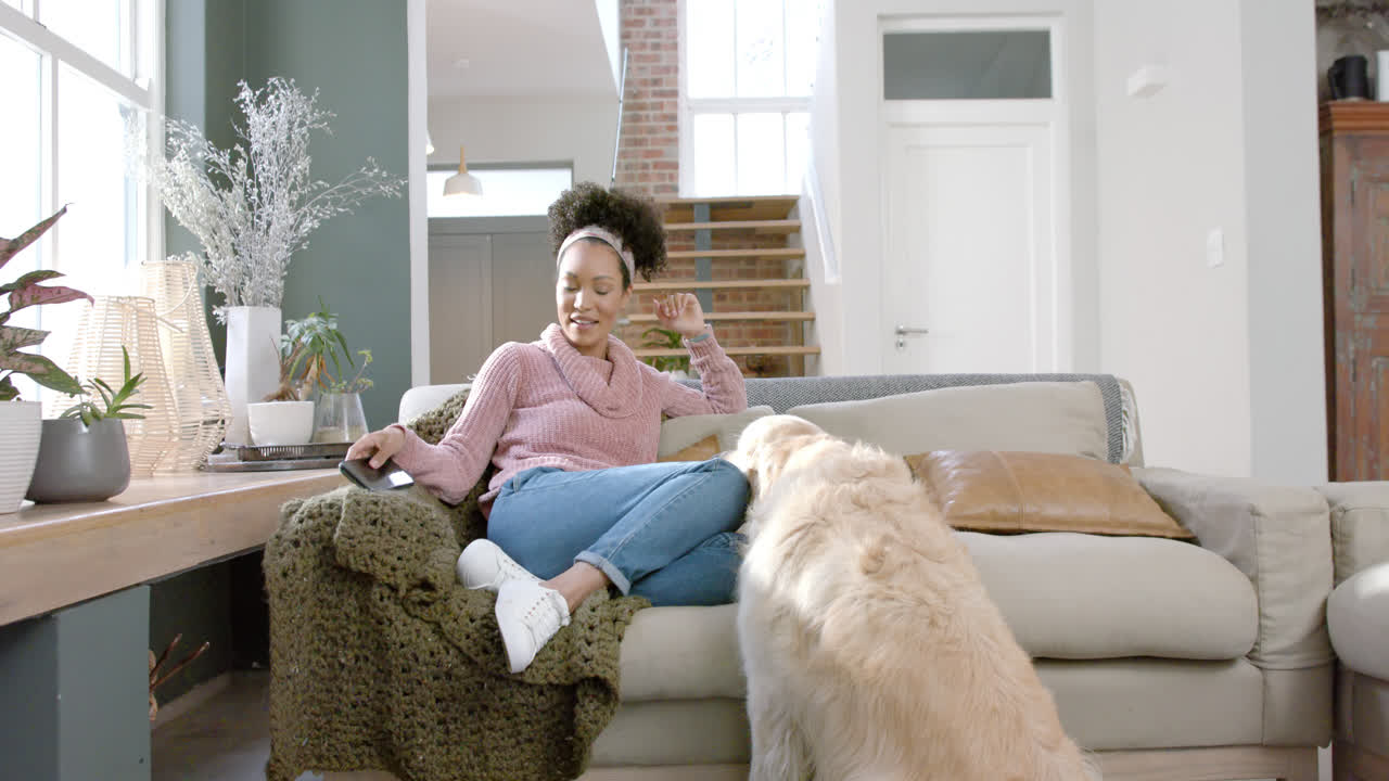 Happy biracial woman petting golden retriever dog and using smartphone at home, slow motion
