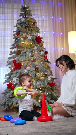 caucasian mother and baby playing happily near christmas tree. little toddler boy constructs a pyramid. vertical video.