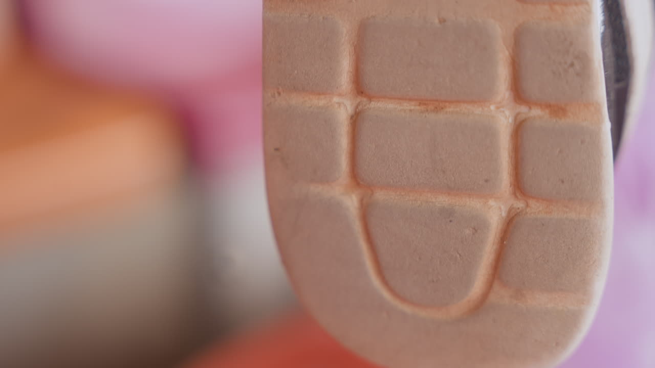 Primo piano del battistrada di una scarpa da ginnastica con suola in gomma morbida a griglia. Lo sfondo rosa sfocato evoca una giocosa scena di sala giochi per bambini, con suola esterna usurata e sottili segni di usura. La macrofotografia cattura la texture.