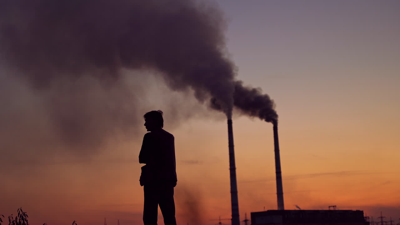 Massive dark smoke going from pipes at sunset. Silhouette of a man standing against the evening sky with dirty dense emissions in the air. Air pollution.