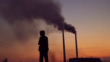 massive dark smoke going from pipes at sunset. silhouette of a man standing against the evening sky with dirty dense emissions in the air. air pollution.