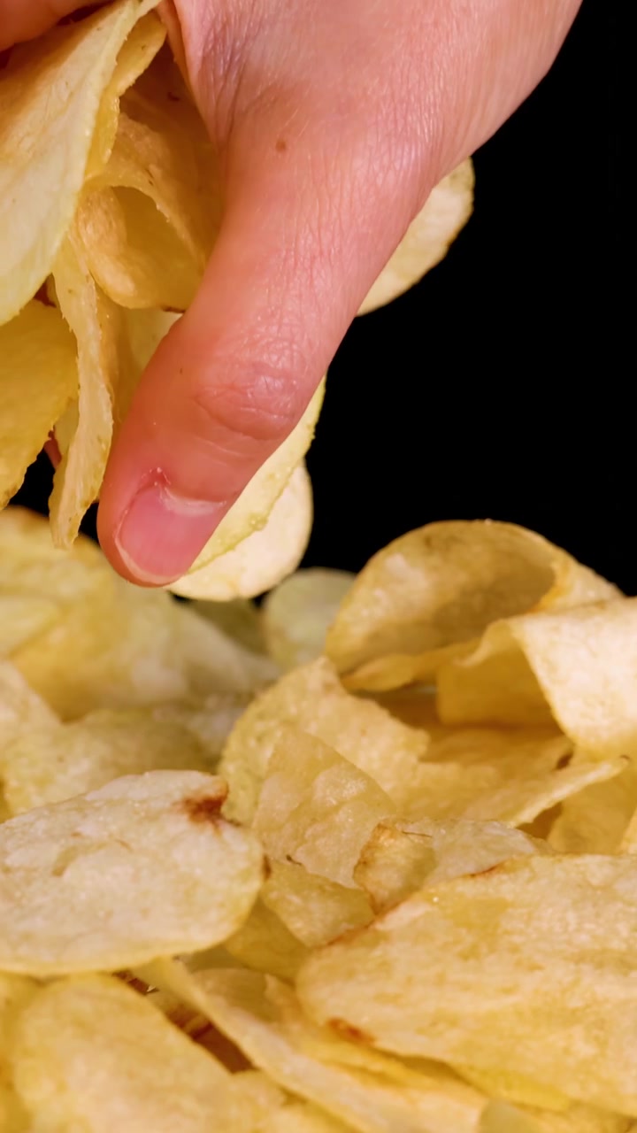 Mão humana pegando batatas fritas de uma pilha, em primeiro plano, iluminação de estúdio, fundo preto.