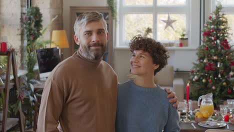 couple looks at the camera and smiles in living room with table and christmas decorations