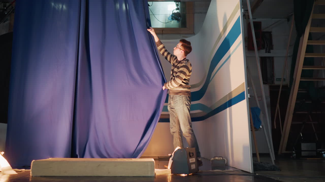 Male operator pulls blue curtain inside television studio set during production setup, floor lights flare below, overhead rigging visible, revealing striped backdrop ready for live news recording