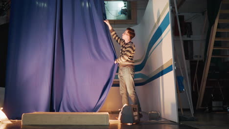 male operator pulls blue curtain inside television studio set during production setup, floor lights flare below, overhead rigging visible, revealing striped backdrop ready for live news recording