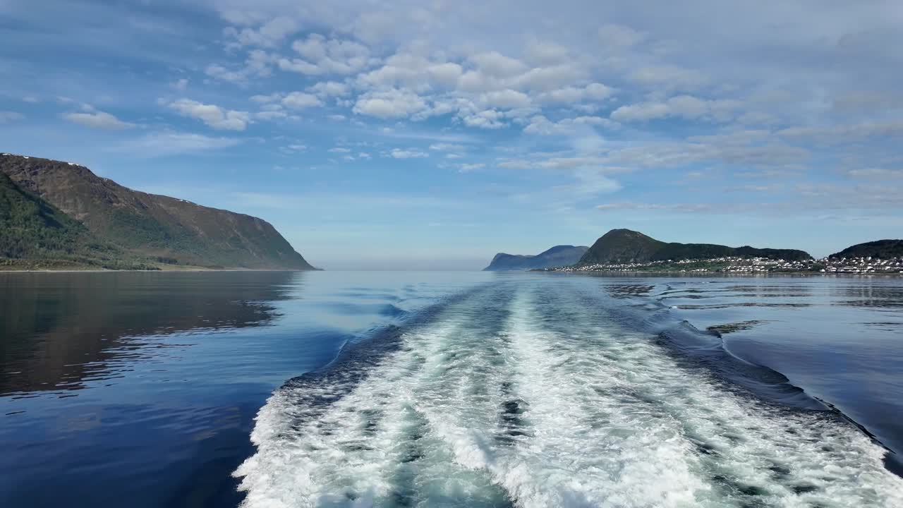 Fantastisk bakåtblick från en rörlig båt som lämnar Ålesund, Norge, med ett vitt skummande kölvatten som sträcker sig över lugna fjordvatten, flankerade av dramatiska berg och blå himmel.