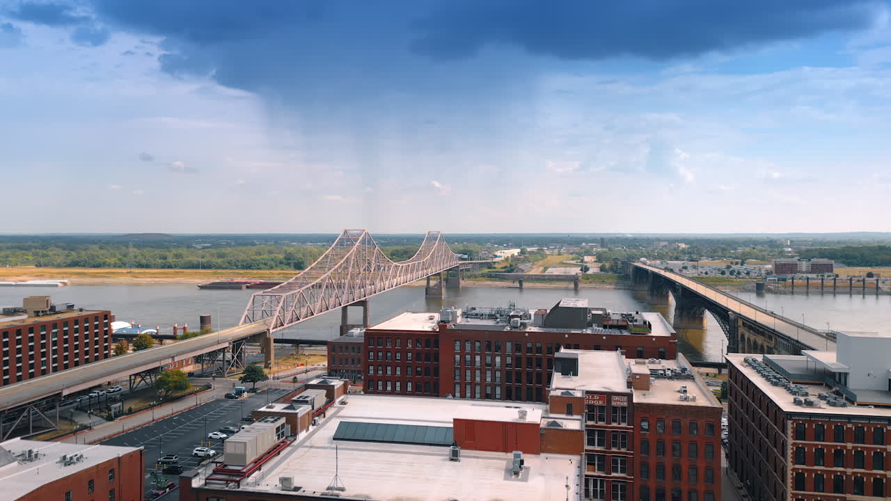 Saint Louis USA, 14 August 2025: Rising above the multi-storied red-brick buildings at the waterfront of the Mississippi River in St. Louis, Missouri, USA. Revealing view on the bridges across the waterscape. Rainy cloud in the sky above