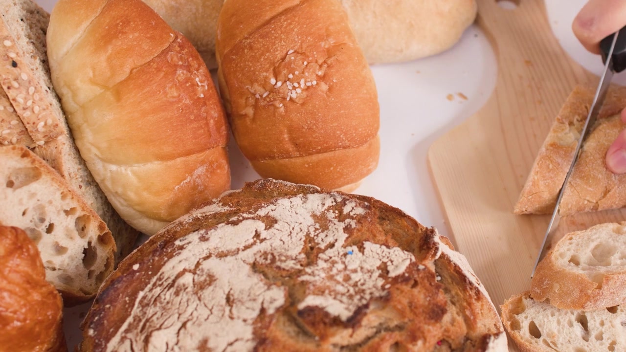 Close-up of hands cutting crusty bread on a wooden board amidst various artisanal pastries.