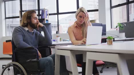 Disabled-caucasian-businessman-sitting-on-wheelchair-using-laptop-talking-to-coworker-in-office