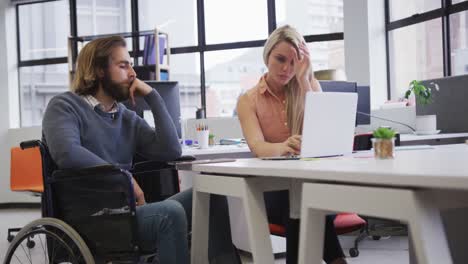 disabled caucasian businessman sitting on wheelchair using laptop talking to coworker in office