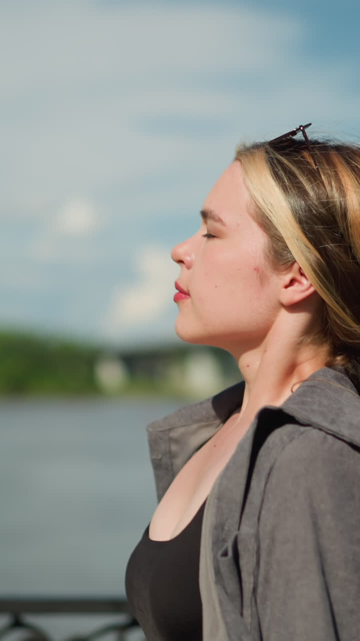 dama de camiseta gris y camiseta negra camina cerca de la orilla del río, pasando por una valla metálica, el fondo presenta una vista borrosa del puente y el agua mientras mira hacia el río y toma una respiración profunda