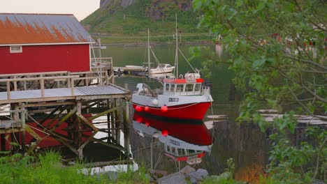 A-red-fishing-boat-docked-near-a-rustic-red-boathouse-in-Reine,-Lofoten-Islands,-Norway