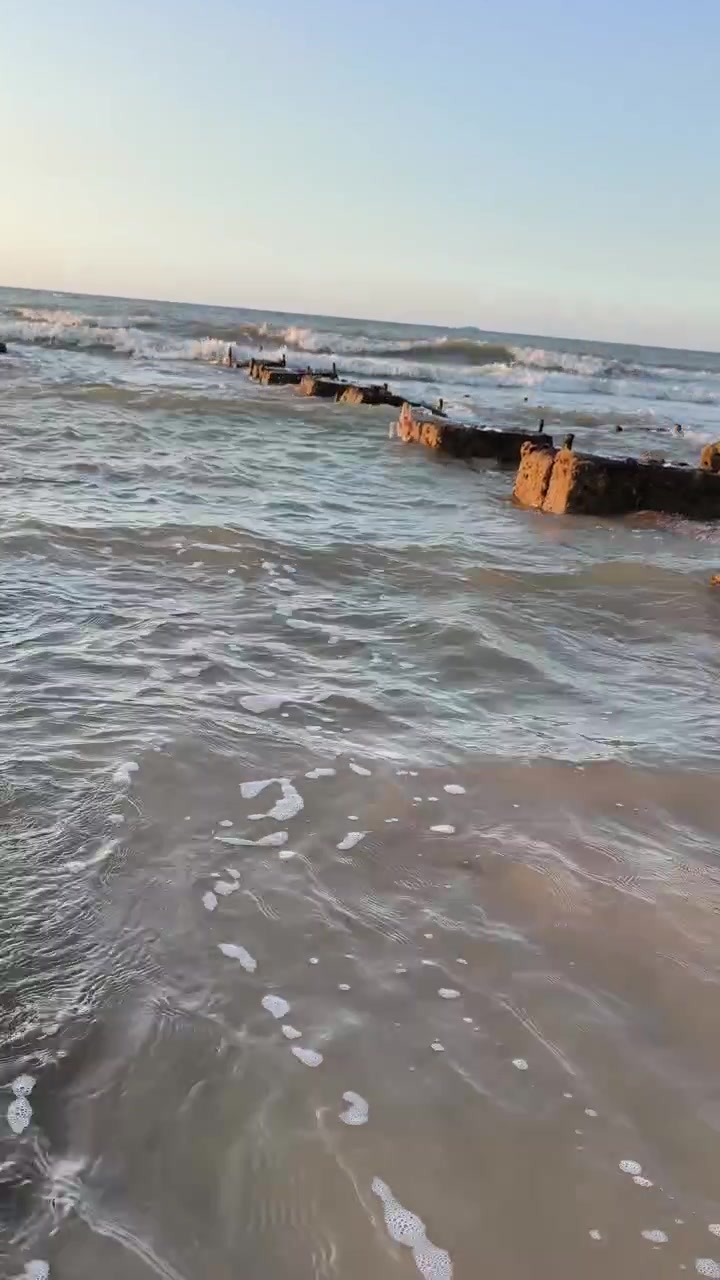 Ondas do oceano quebrando contra ruínas de um antigo píer de concreto ao pôr do sol.