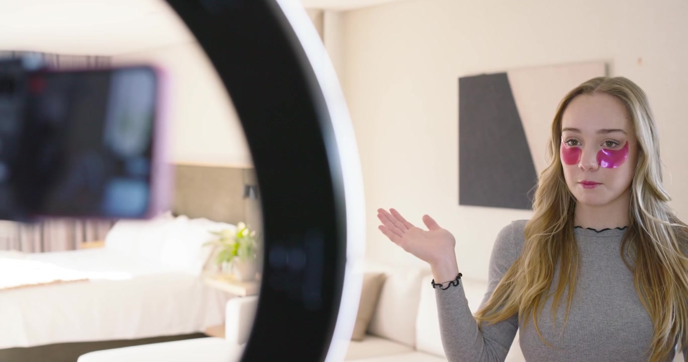 Donna adulta che reagisce a una ring light, pulendosi la zona sotto gli occhi con un dischetto e applicando patch rosa.