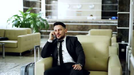 pan shot of confident businessman talking mobile phone while sits on armchair in luxury hotel with luggage near him. travel, business and people concept