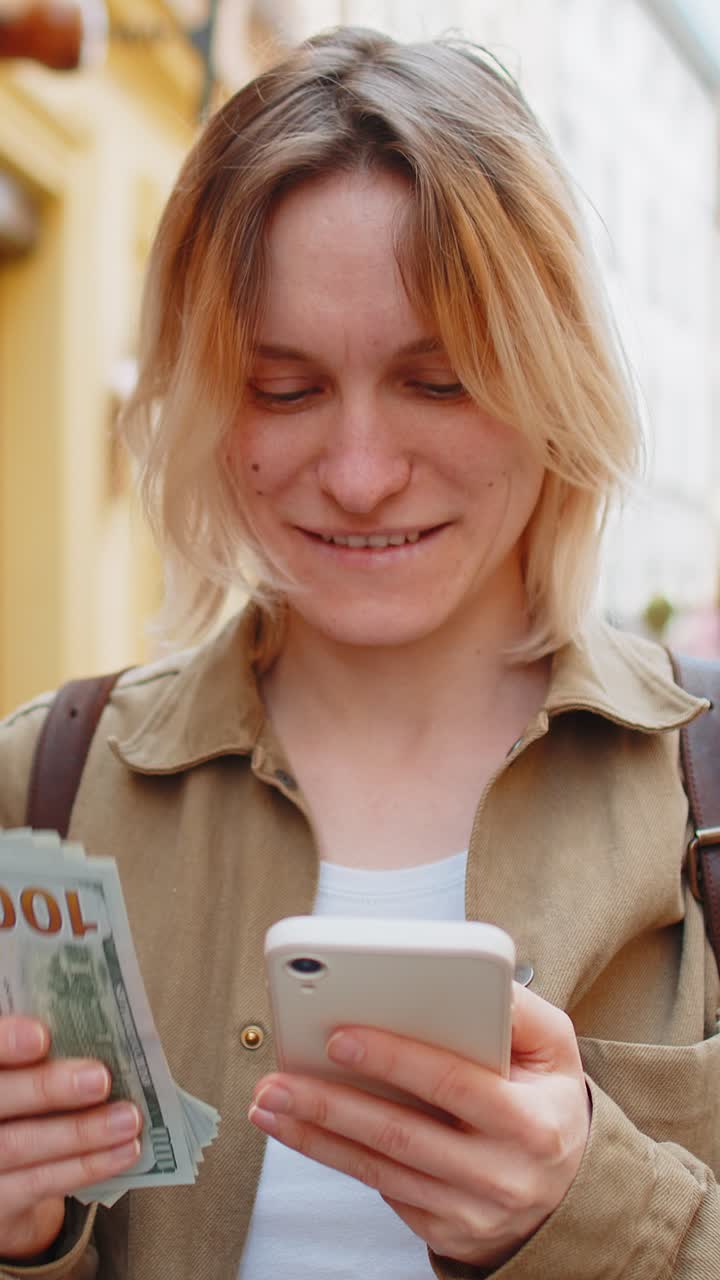Donna che conta soldi in dollari in contanti usando un'app di calcolatrice per smartphone soddisfatta dello stipendio in una strada della città