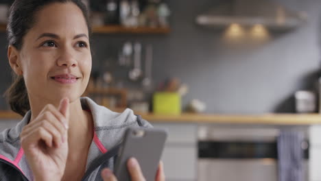 Bonita-Mujer-De-Raza-Mixta-Usando-Un-Teléfono-Inteligente-Con-Pantalla-Táctil-En-Casa-En-La-Cocina-Usando-Ropa-Deportiva-Bebiendo-Café-Sonriendo