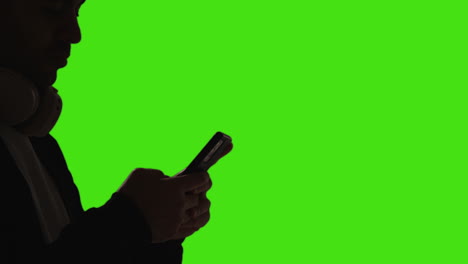 close up of man wearing wireless headphones text messaging on mobile phone against green screen with low key lighting