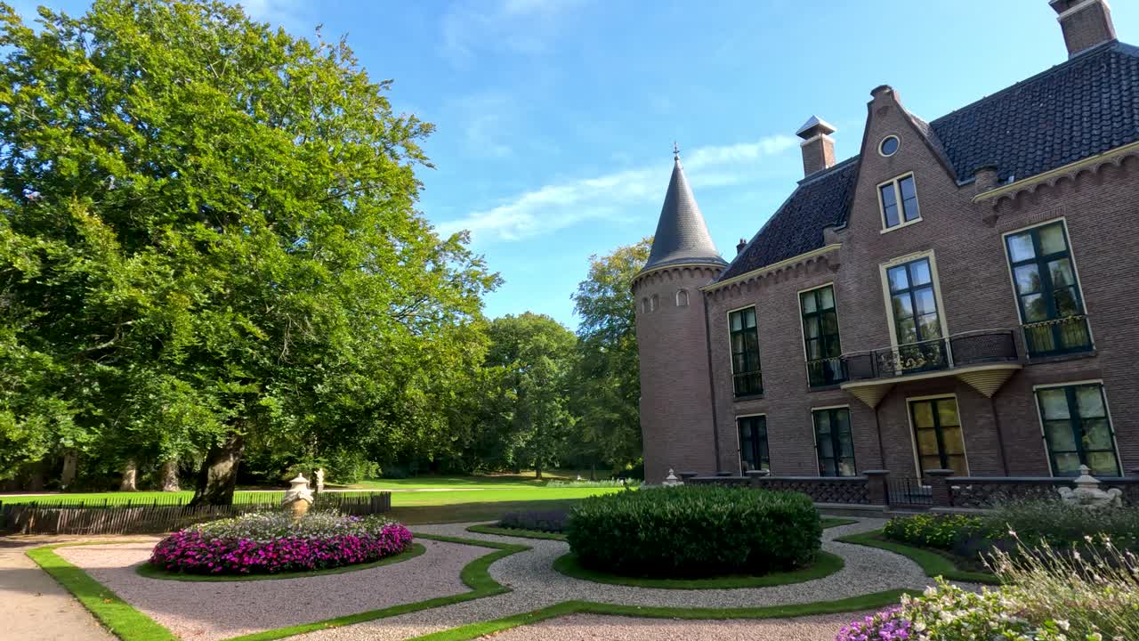 Daytime camera pan reveals a historic brick castle with turrets, manicured gardens, and vibrant flowerbeds under clear blue skies in Lisse, Netherlands