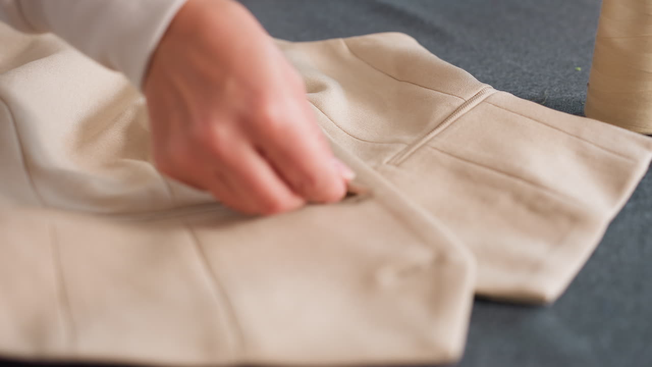 Seamstress hand positioning brown button into opening on beige tailored jacket laid on worktable, planning fastening alignment during garment finishing process inside fashion studio