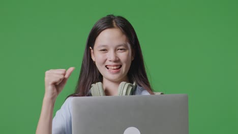 배을 가진 아시아 십대 소녀 학생이 녹색 스크린 배경 스튜디오에 서서 노트북을 사용하고 축하합니다.