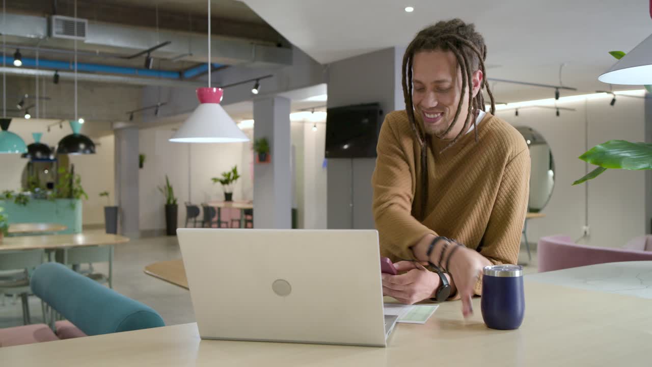 African American man tapping phone reading texts, sipping tumbler checking laptop in workspace