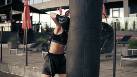 Attractive-athletic-female-boxer-in-gloves-kicking-a-punching-bag.-Workout-outside.-Female-boxer-training.-Self-defence-concept