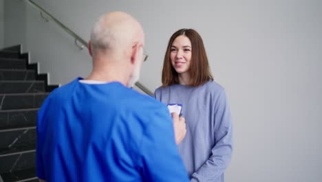 sobre el hombro una confiada chica morena con una chaqueta azul se comunica con un médico experimentado con cabello gris en gafas y un uniforme azul que tiene una tableta con diagnósticos y consejos en sus manos en una clínica moderna