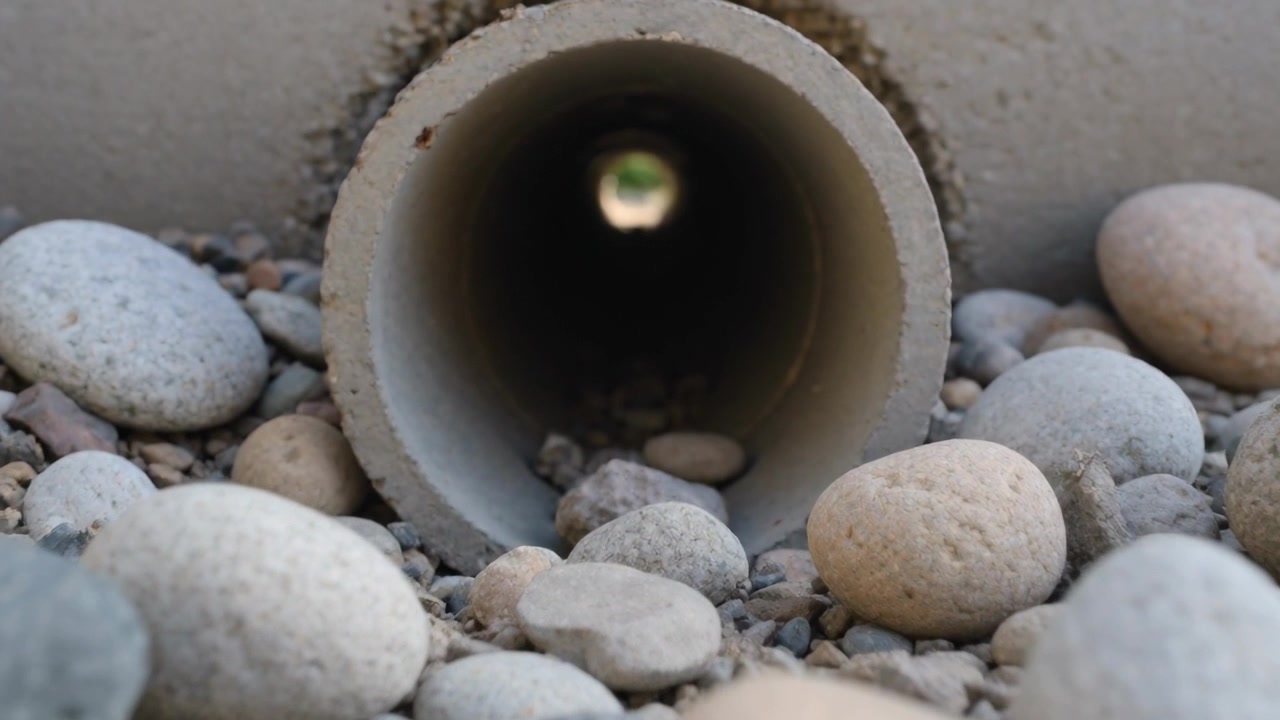 Moving camera revealing concrete pipe at ground level among pebbles, showing distant greenery