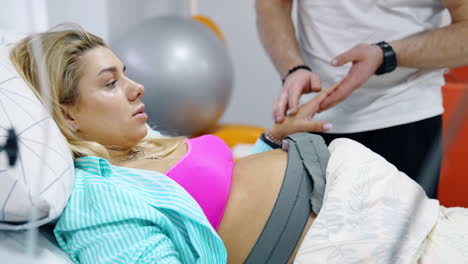beautiful blonde pregnant woman lies in hospital bed. belt from medical equipment is on woman's belly. man standing beside hold his wife by the hand.