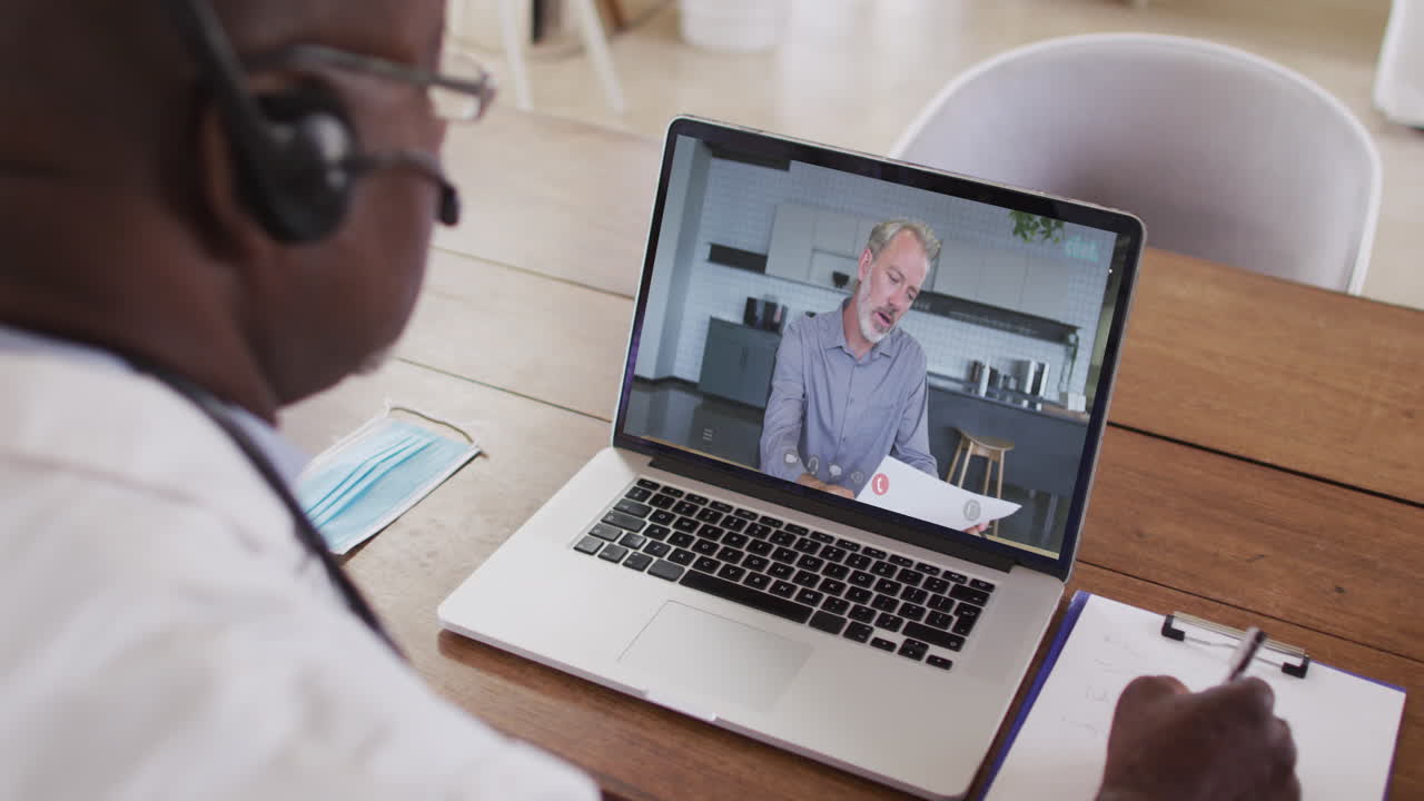 African american male doctor wearing phone headset taking notes while having a video call on laptop