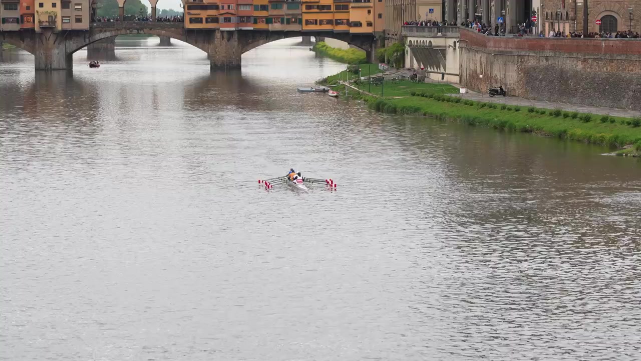 Wide shot of rowers gliding on the Arno River beneath the iconic Ponte Vecchio in Florence, Italy. Overcast daylight enhances historic architecture, vibrant water activity, and scenic city life.