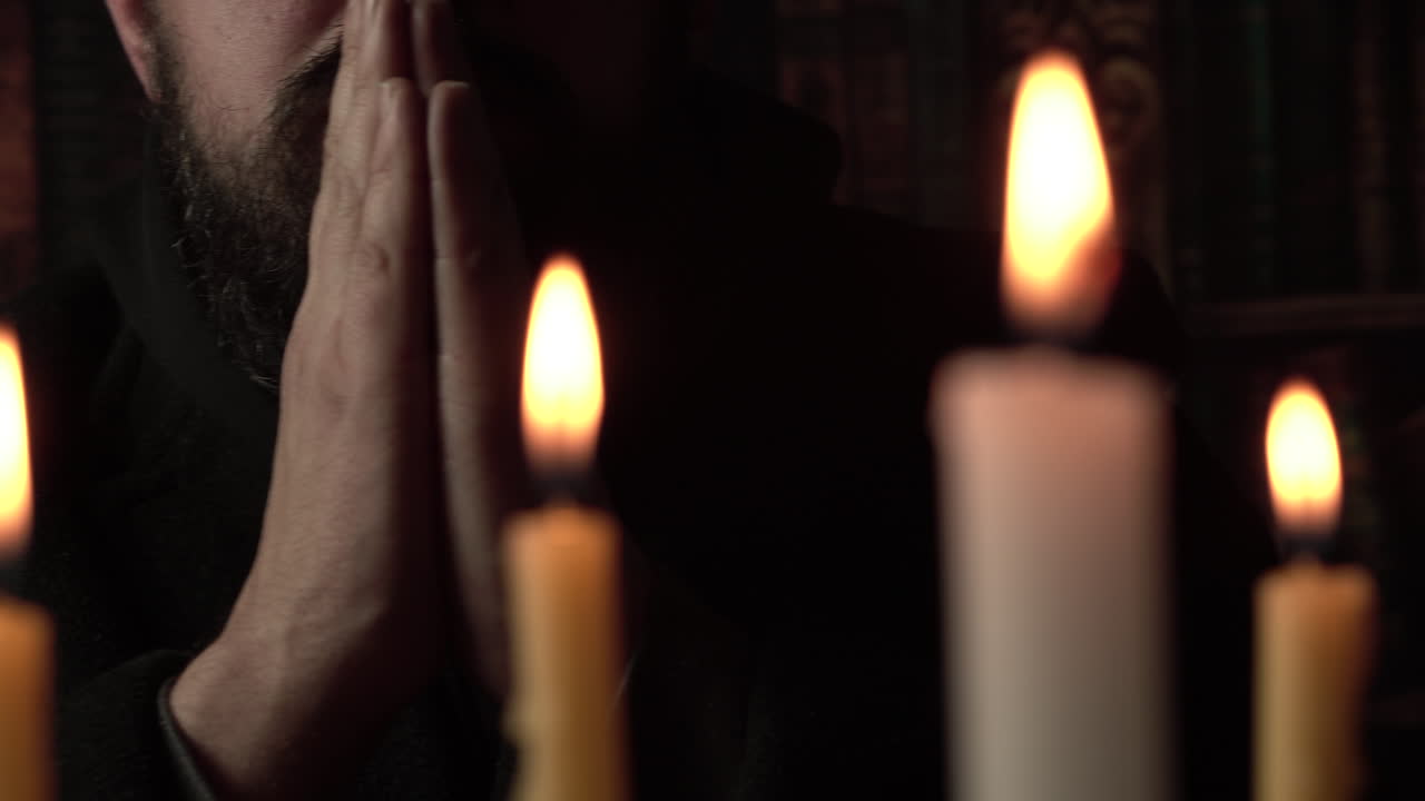 Uomo con le mani giunte che prega Dio davanti a delle candele, primo piano.