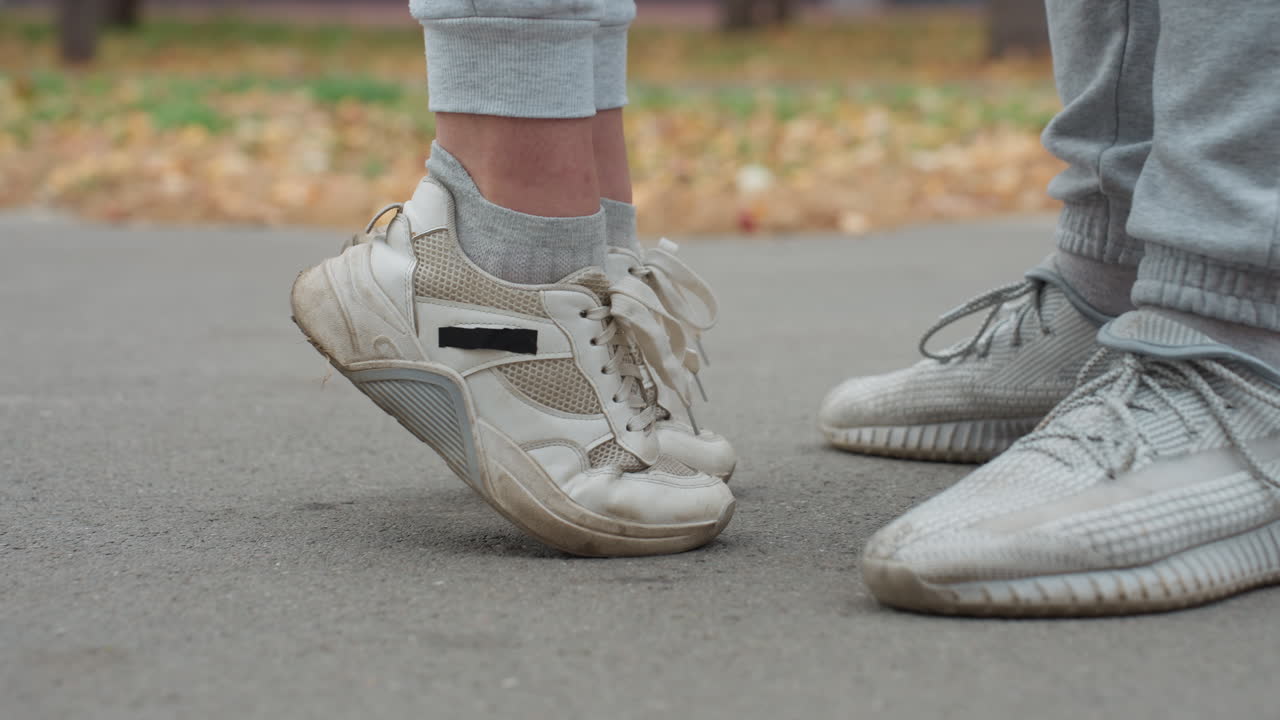 Leg view of two people standing close together in joggers and sneakers on paved path with blurred background featuring dry autumn foliage and tree