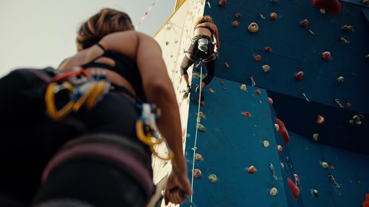 desde abajo, una chica rubia con un uniforme de verano deportivo negro con el equipo y el seguro necesarios sube a una pared de escalada azul, y una chica con un peinado de bob en un uniforme negro de verano la asegura y sostiene la cuerda necesaria para apoyar a su amiga.