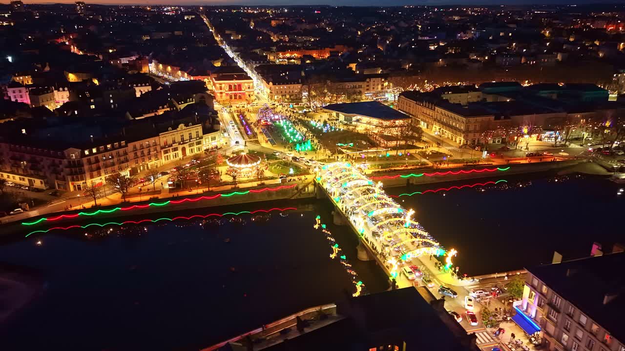 Drone panning right across Laval at night, showing the illuminated Aristide Briand Bridge, the festive carousel, the vibrant Christmas displays and the reflections along the Mayenne River
