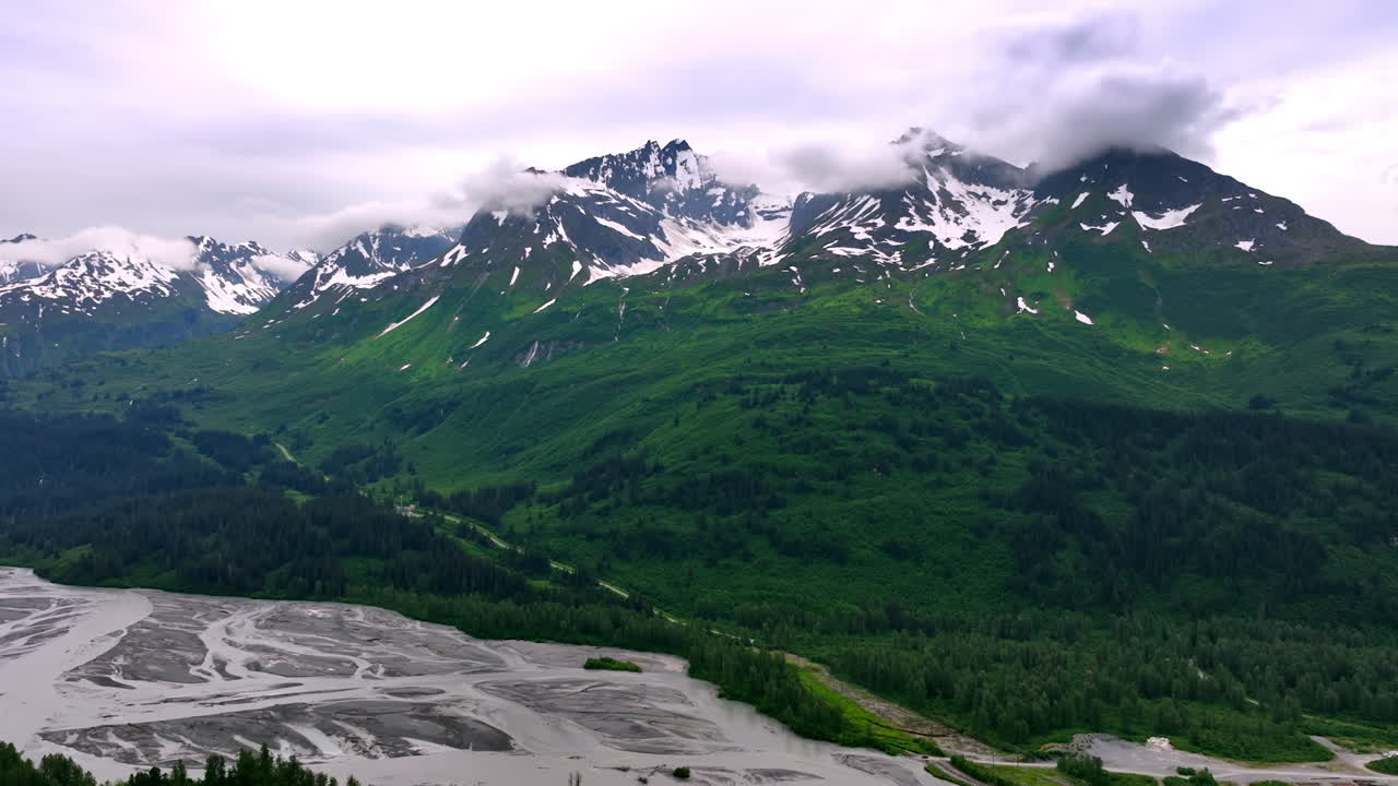 Clouds hide the peaks of the mountains covered with snow at tops and greenery at the foot. Branching river flows in the valley. Alaska, USA. Aerial view