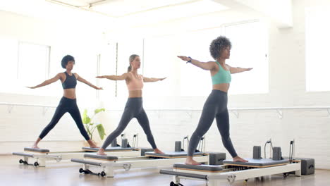 three women practicing yoga on pilates reformers in bright studio