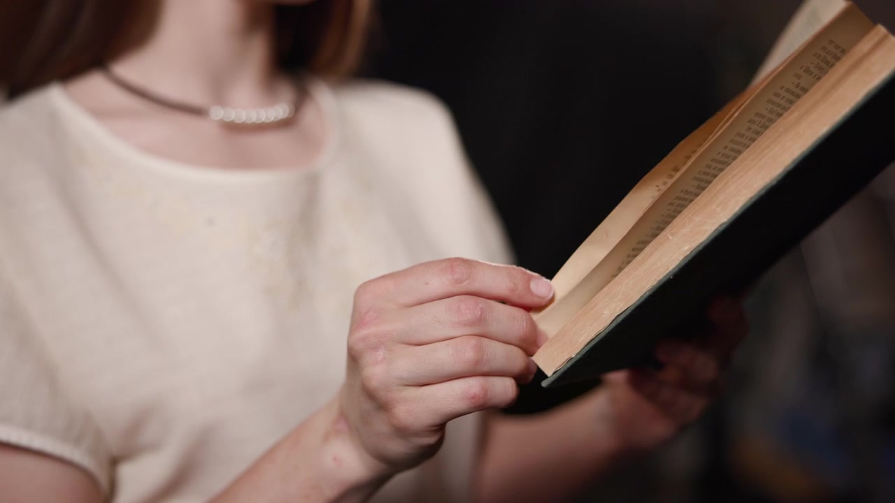 Una giovane donna con un top chiaro tiene un libro aperto vicino al petto in un ambiente interno con luce soffusa, con uno sfondo scuro e sfocato e una messa a fuoco ridotta su di lei.