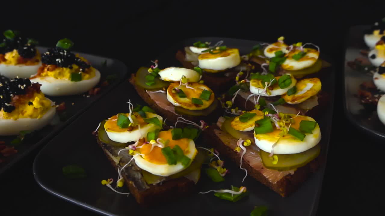 Délicieux œufs mimosa au caviar noir et sandwichs au hareng, cornichons et pousses, disposés sur des assiettes noires pour une célébration, une fête ou un événement traiteur sur fond sombre.