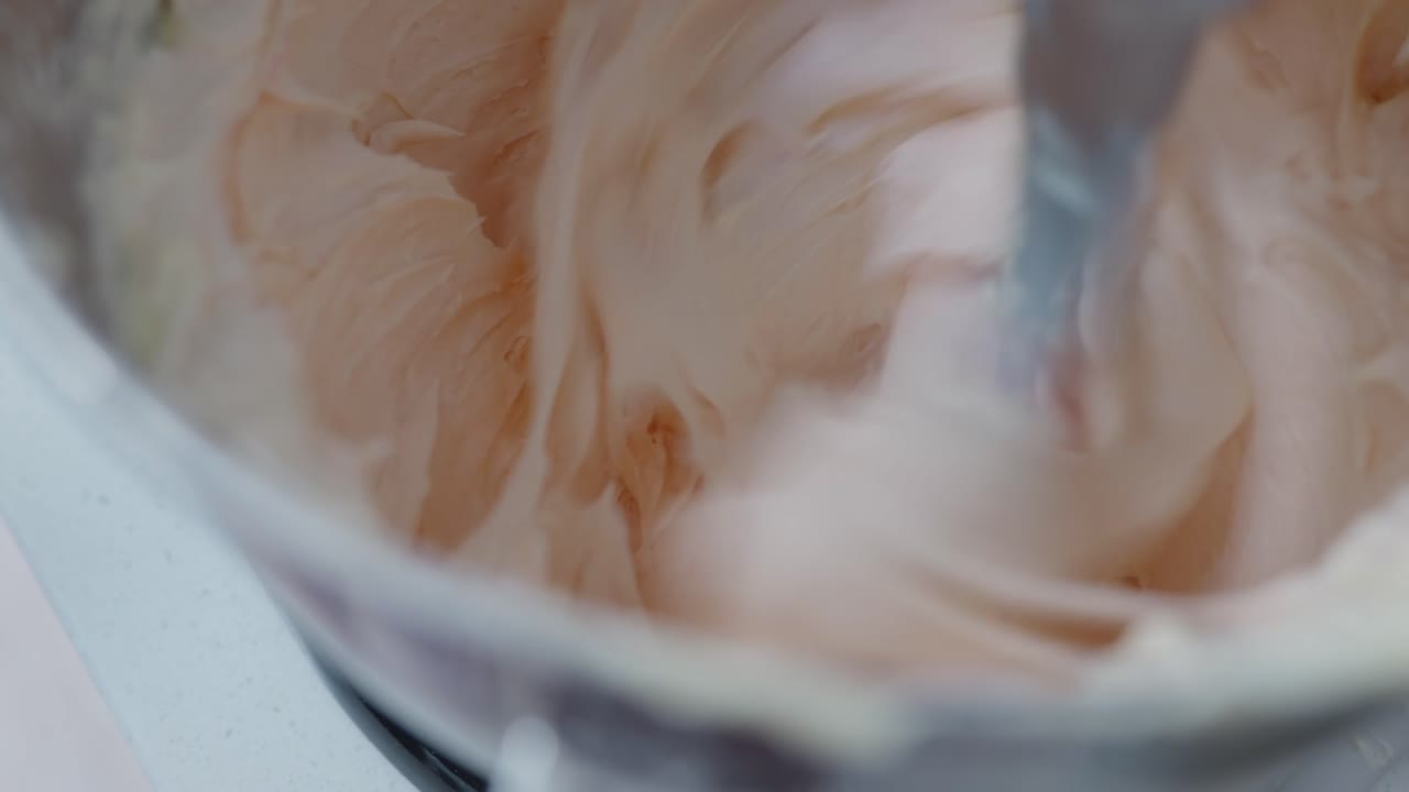 Close up, A cook drops food colouring into pink buttercream icing whipping in a stand mixer bowl on high speed
