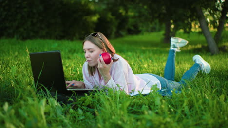 young woman lying on grass using laptop and eating an apple surrounded by lush greenery and trees, creating a peaceful outdoor work environment with a natural and vibrant backdrop