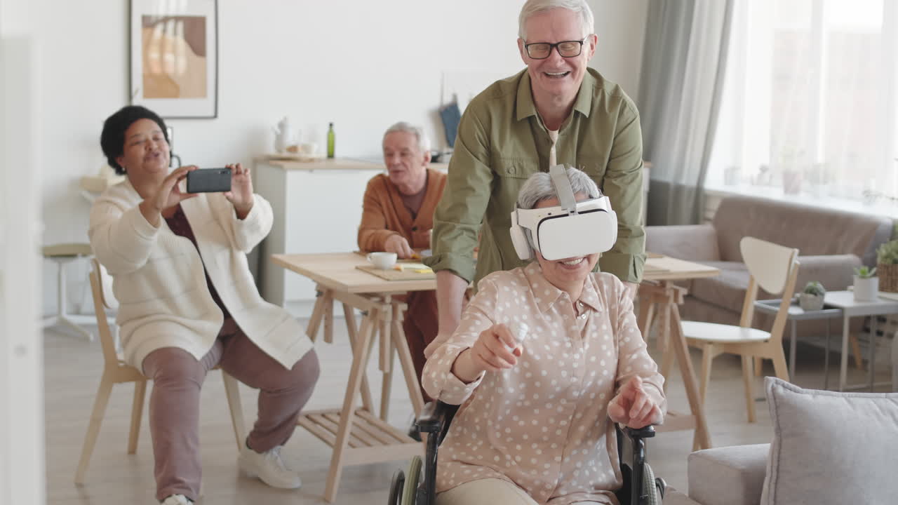 Plano medio largo de una mujer feliz con discapacidad usando gafas de realidad virtual con auriculares, su amiga caucásica mayor moviendo la silla de ruedas de lado, una mujer filmándolos con un smartphone y sonriendo de fondo.