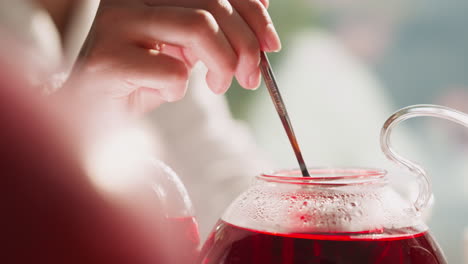 woman stirs tea in glass teapot with spoon closeup. lady prepares hot herbal drink for breakfast at table. process of brewing aromatic hibiscus beverage