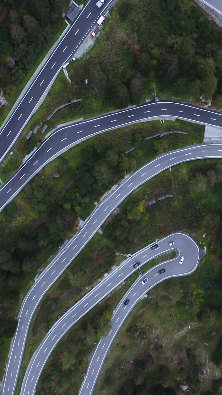Aerial portrait rotating above traffic on a curvy road, in the cloudy alps