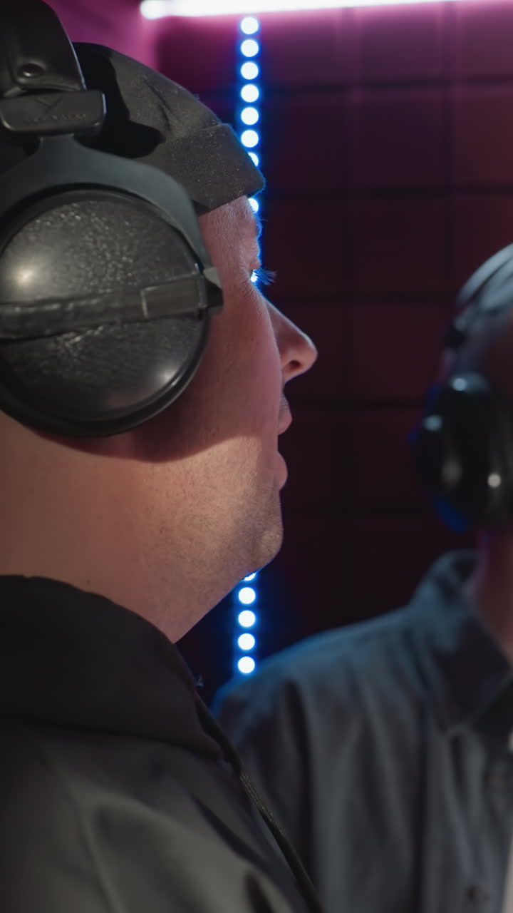 소음 방지 녹음 스튜디오에 있는 두 남자가, 검은 모자와 후디를 입은 한 남자가 다른 남자를 지켜보면서 미소 짓고, 파란 셔츠와 색 하부 셔츠를 입고 열정적으로 노래하고 있다.