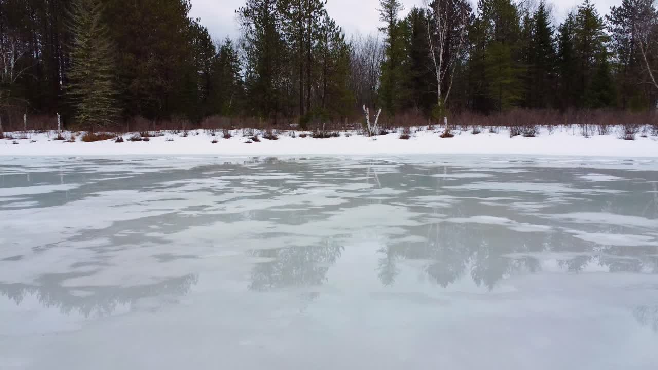 Thin meltwater reflects forest on frozen pond in slow forward glide